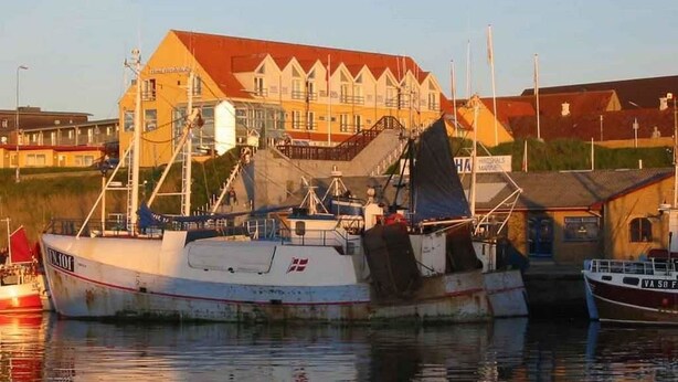Un puerto con una embarcación pesquera grande atracada, un edificio con techo rojo y una lancha cerca.