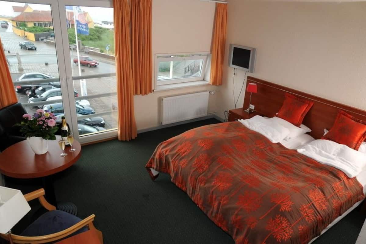 Ein Hotelzimmer mit Bett, Fernseher, Fenster mit Vorhängen, Tisch mit Vase und Blumen sowie Blick auf geparkte Autos vor dem Haus.