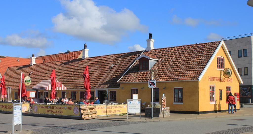 A yellow building with a red-tiled roof, labeled "Hirshals Kro," featuring outdoor seating with red umbrellas.