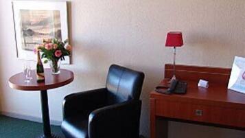 A hotel room with a black leather chair, a small wooden table with a red lamp, a telephone, and a framed picture on the wall.