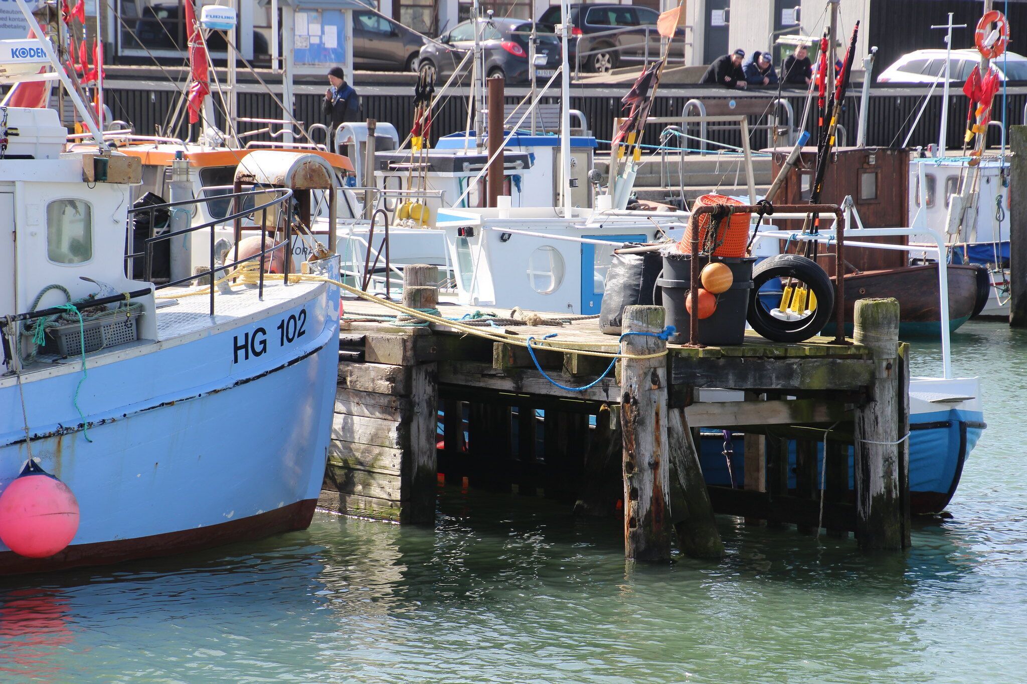 Un puerto con varios barcos amarrados, incluyendo un bote azul con la etiqueta 'HG 102'.