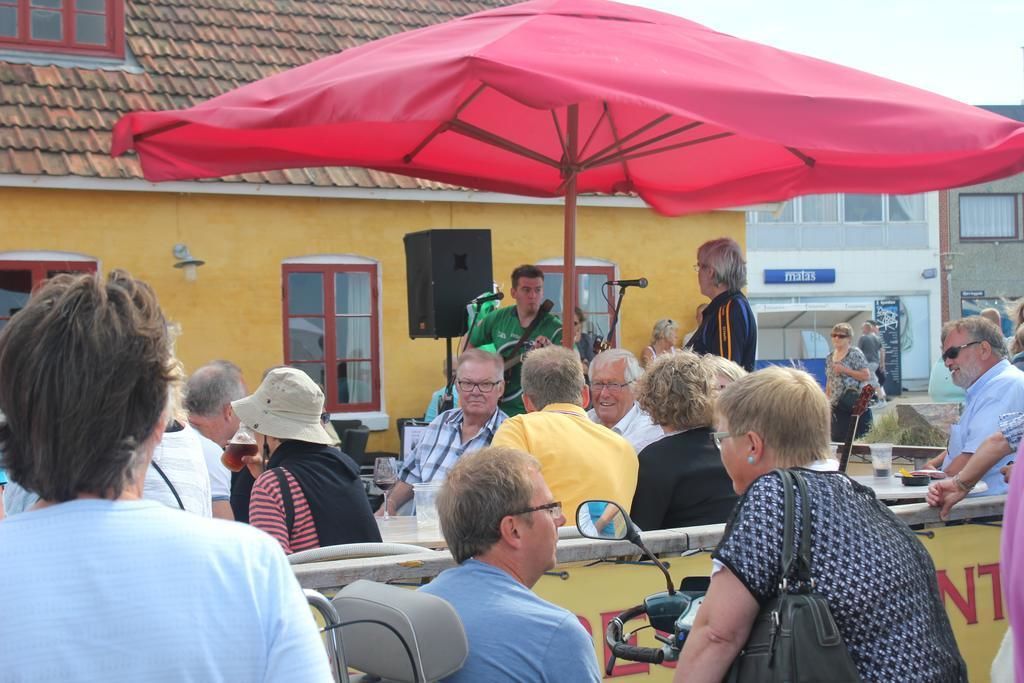 A group of people gathered around a stage with a red umbrella, a yellow building in the background, and a sign that reads "MALAT".
