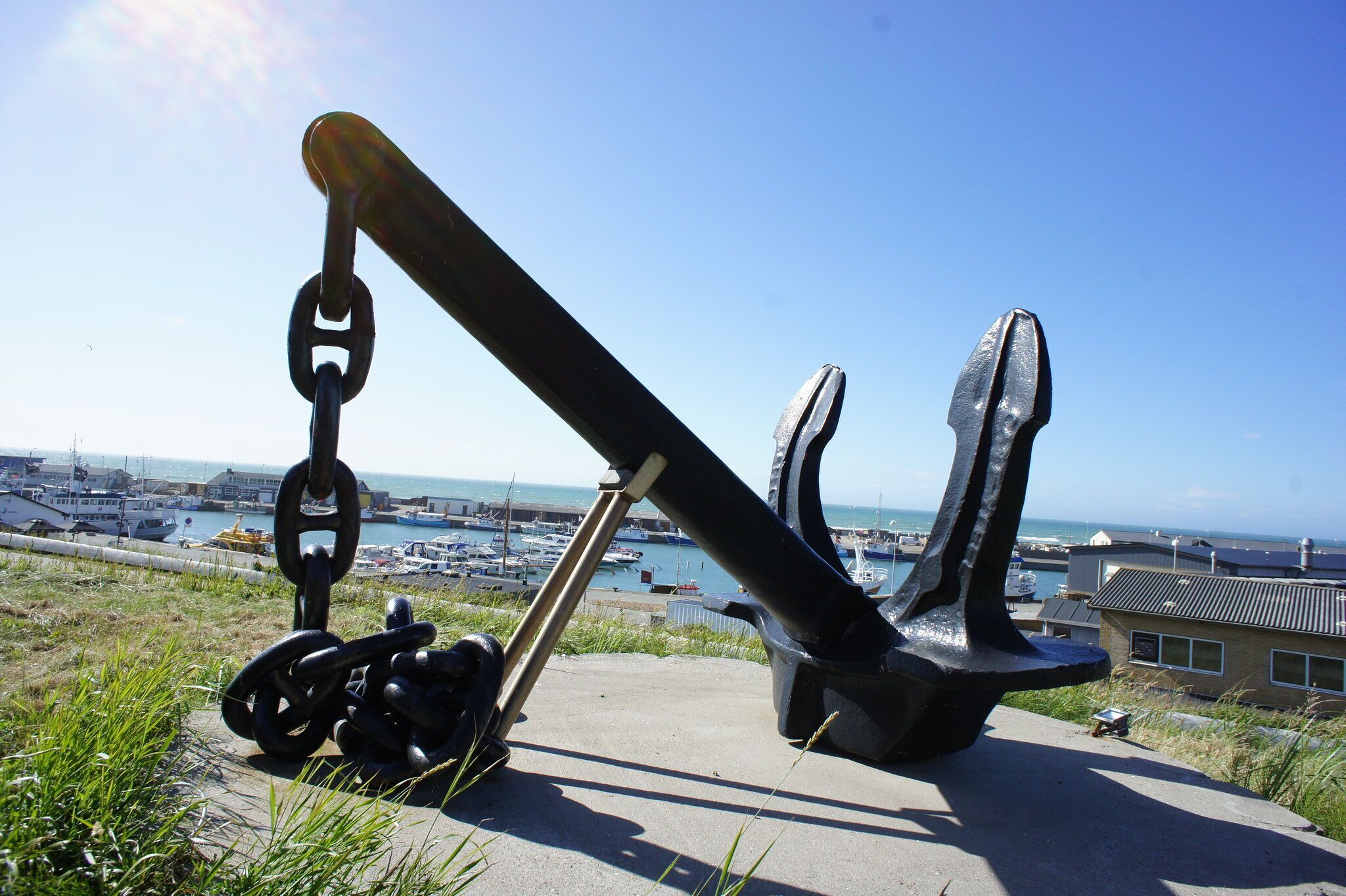 A large anchor sculpture on a concrete platform with a view of a marina and boats in the background.
