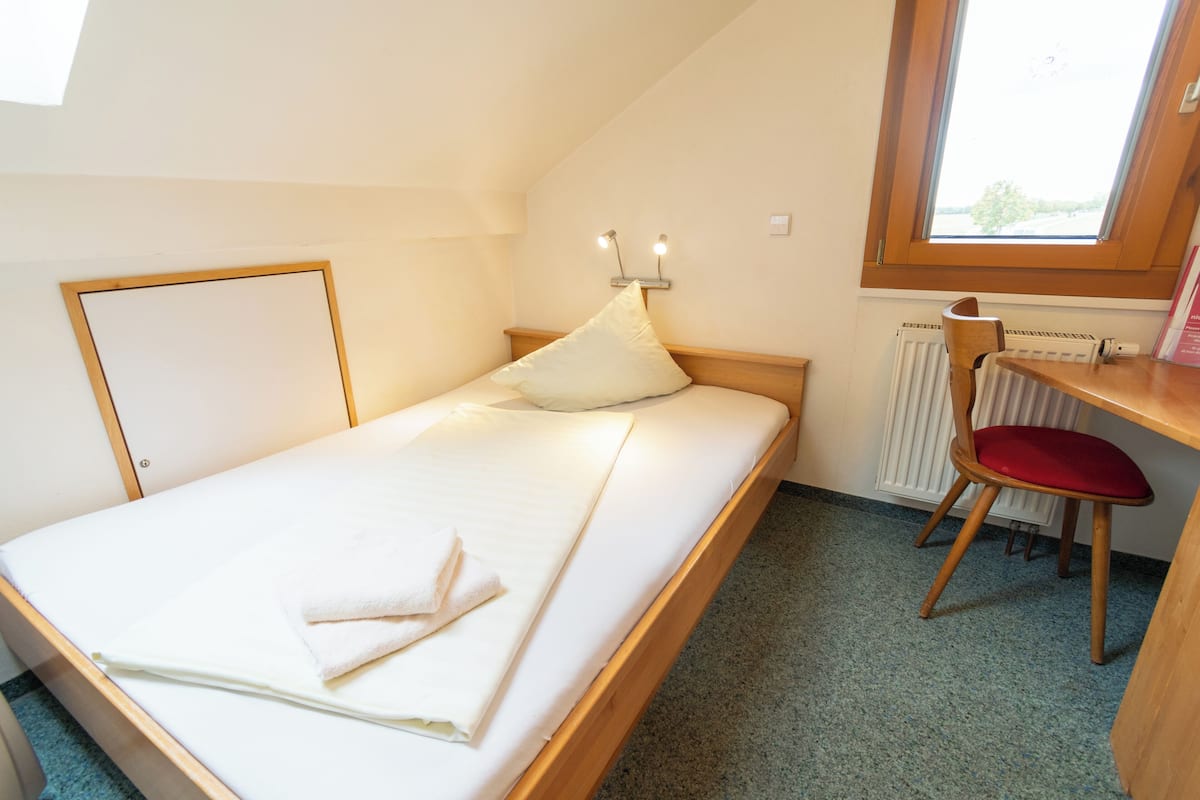 A single bed with a wooden frame, a bedside table with a chair, a window with a view, and a radiator.