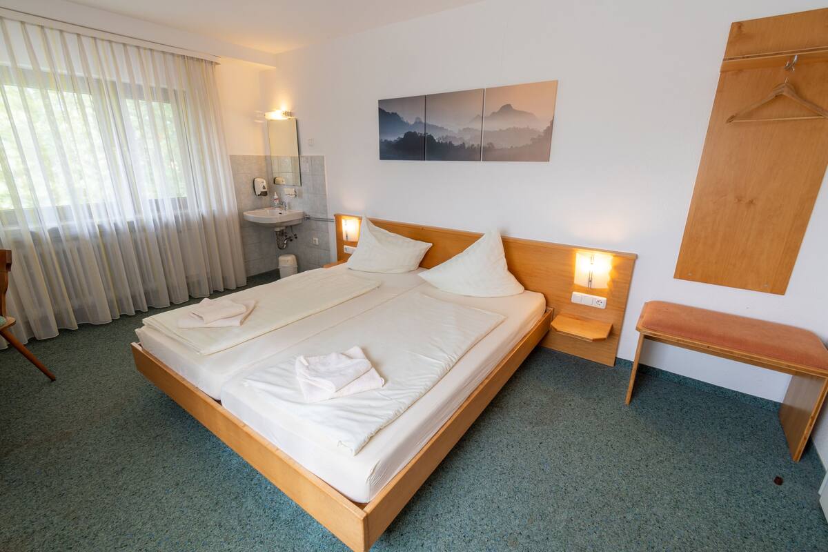 A hotel room with a large bed, bedside tables, a wooden bench, a bathroom with a sink and mirror, and a painting on the wall.