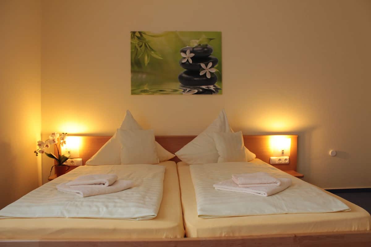 A double bed with white linens and towels, two bedside tables with lamps, and a painting of a zen garden on the wall.