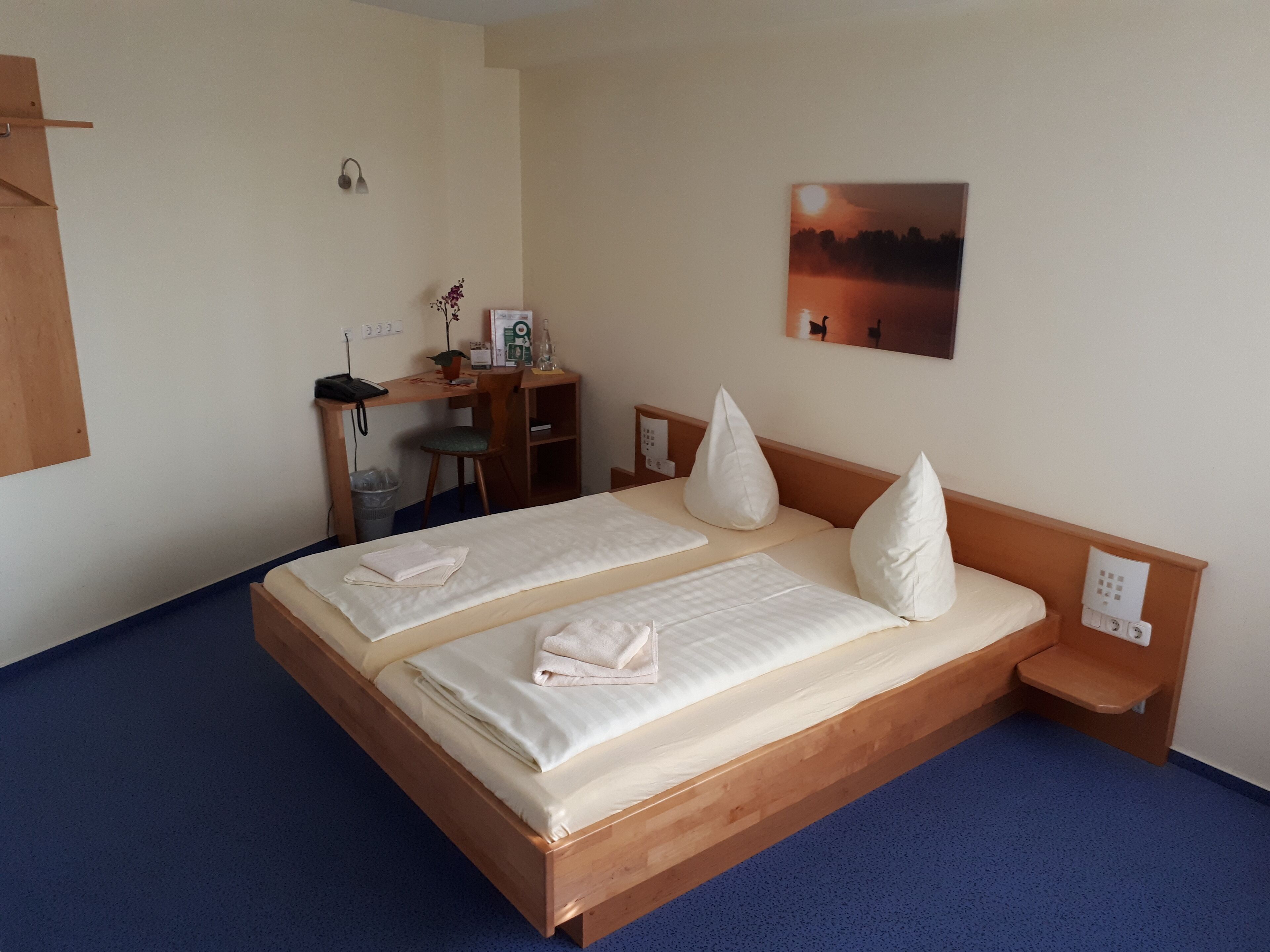 A double bed with white linens and pillows, a desk with a chair, a small table, and a wall-mounted lamp.