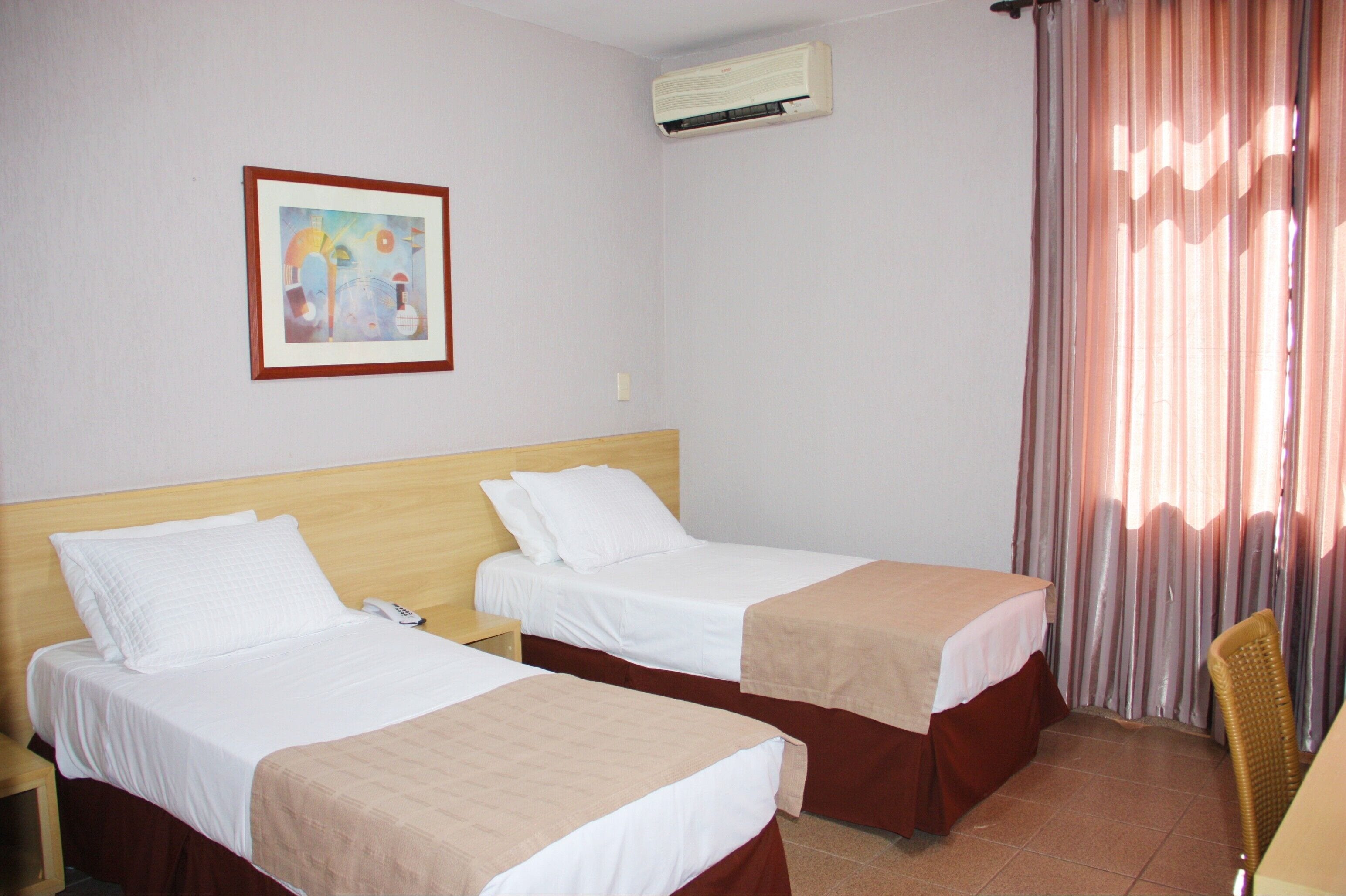 A hotel room with two beds, a painting on the wall, an air conditioning unit, and a window with curtains.