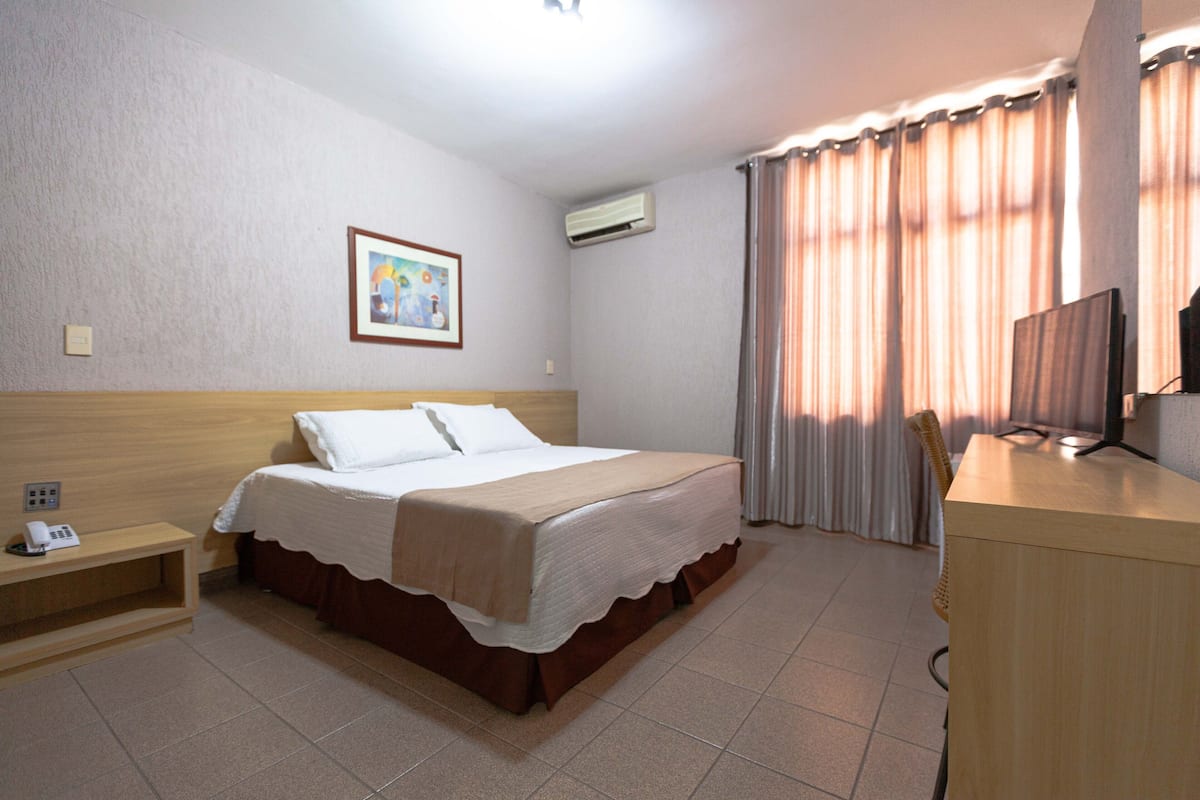 Una habitación de hotel con cama, mesita de noche, escritorio, silla, televisión y cortinas.