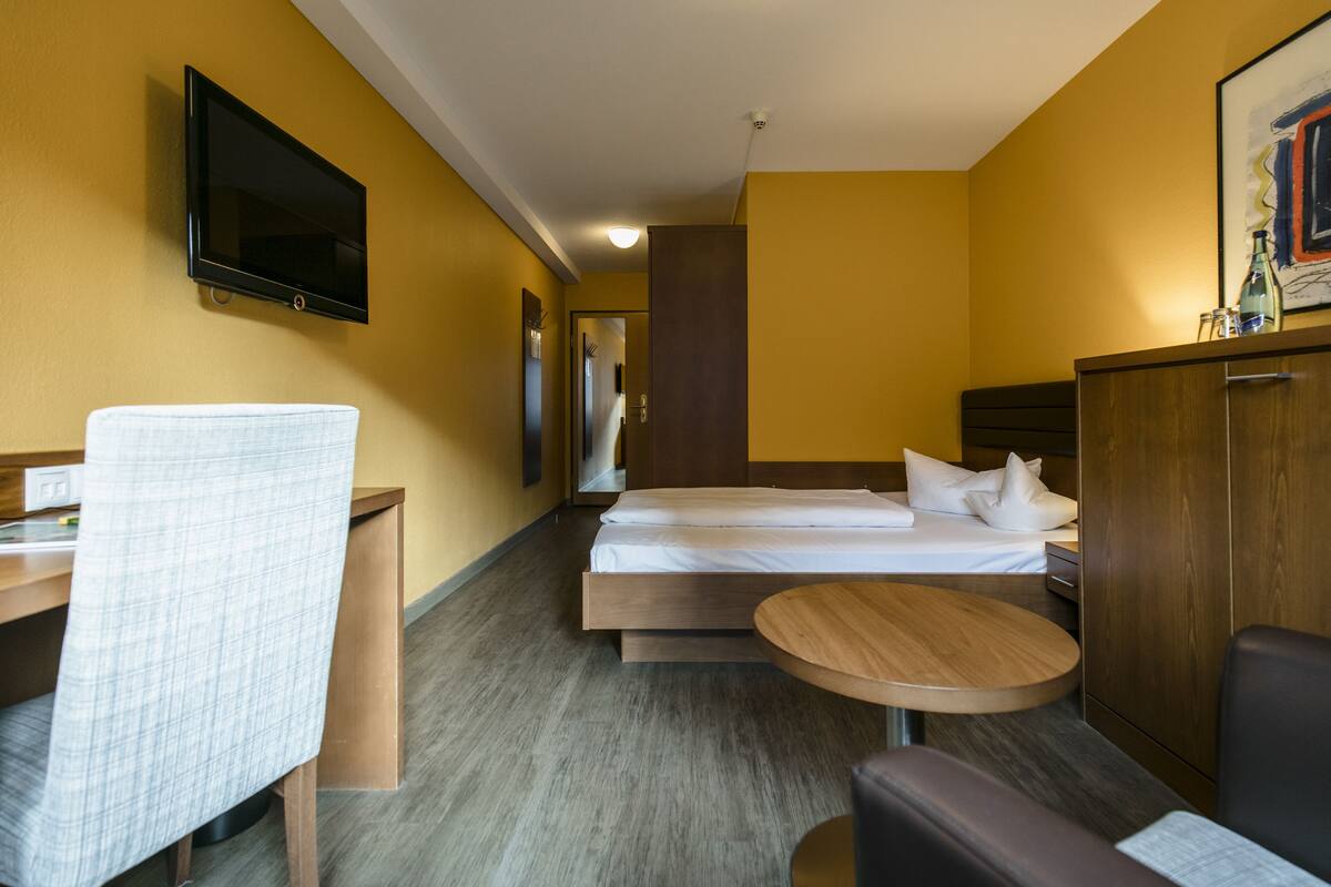 A hotel room with a bed, a desk, a chair, a small table, and a TV mounted on the wall.