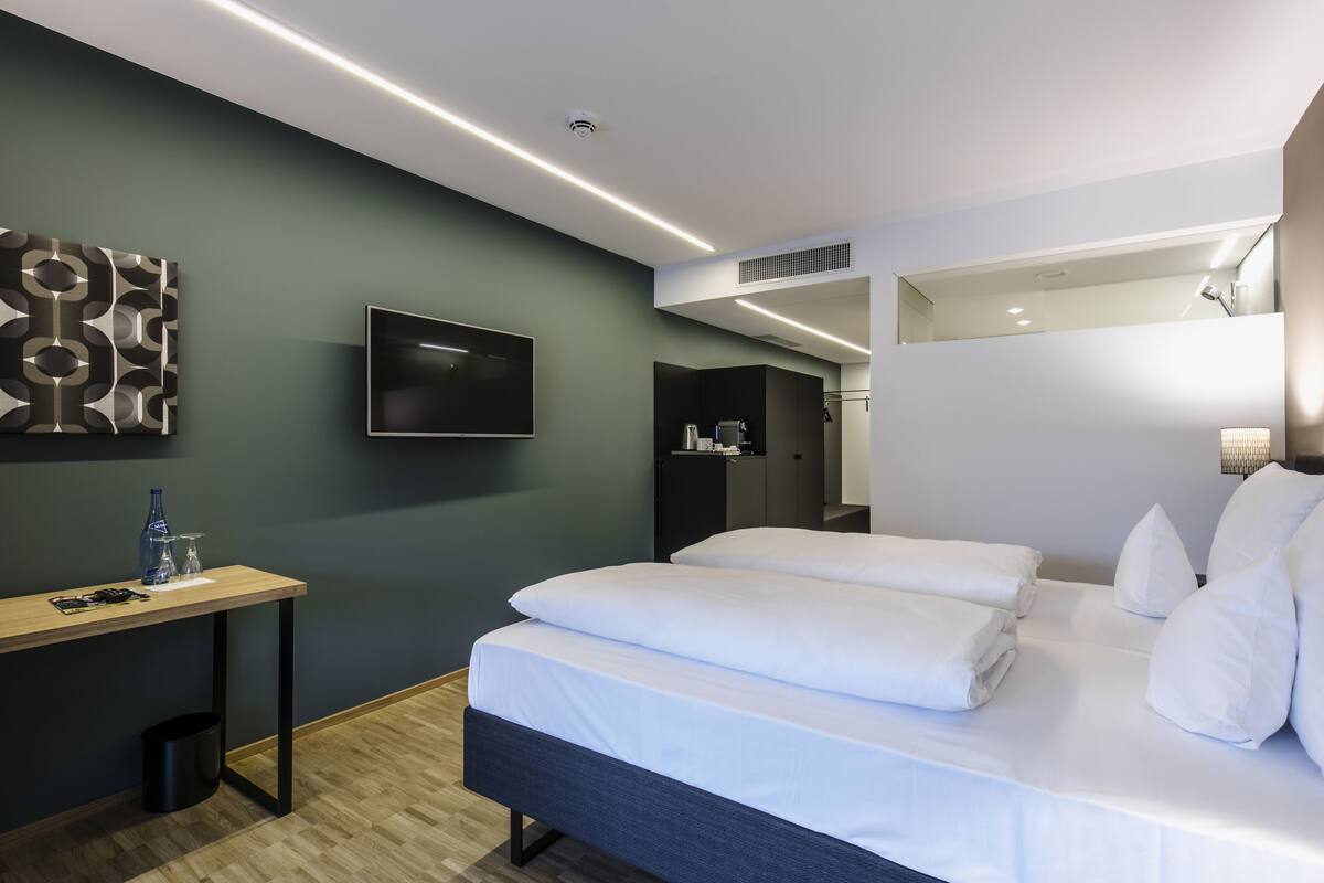 A modern hotel room with a bed, a desk, a TV, and a bathroom visible through an open door.
