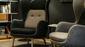 Ein moderner Sessel mit hoher Rückenlehne und einer gepolsterten Oberfläche, platziert in einem Raum mit Bücherregal und einem kleinen Tisch.