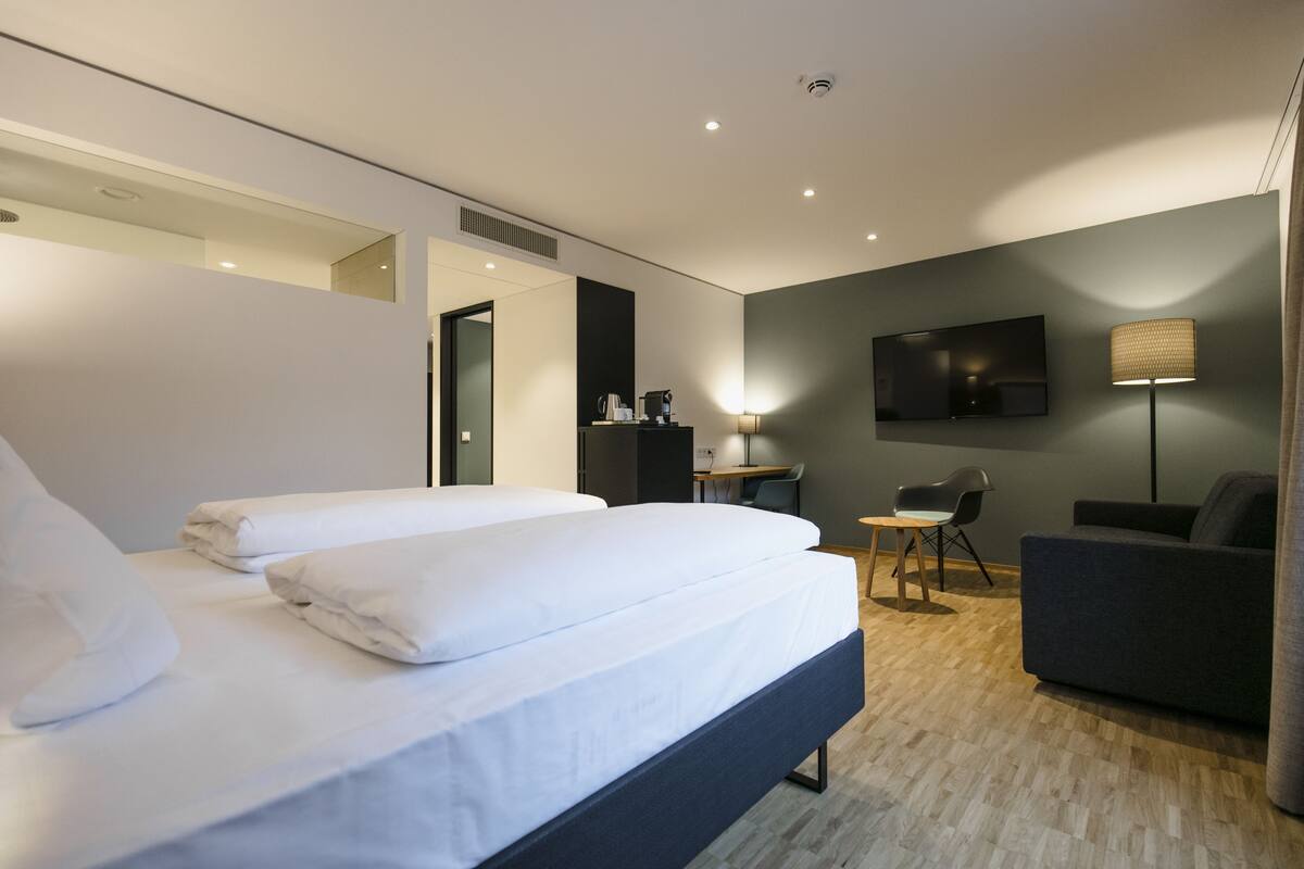 A modern hotel room with a large bed, a desk, a chair, a sofa, and a flat-screen TV mounted on the wall.