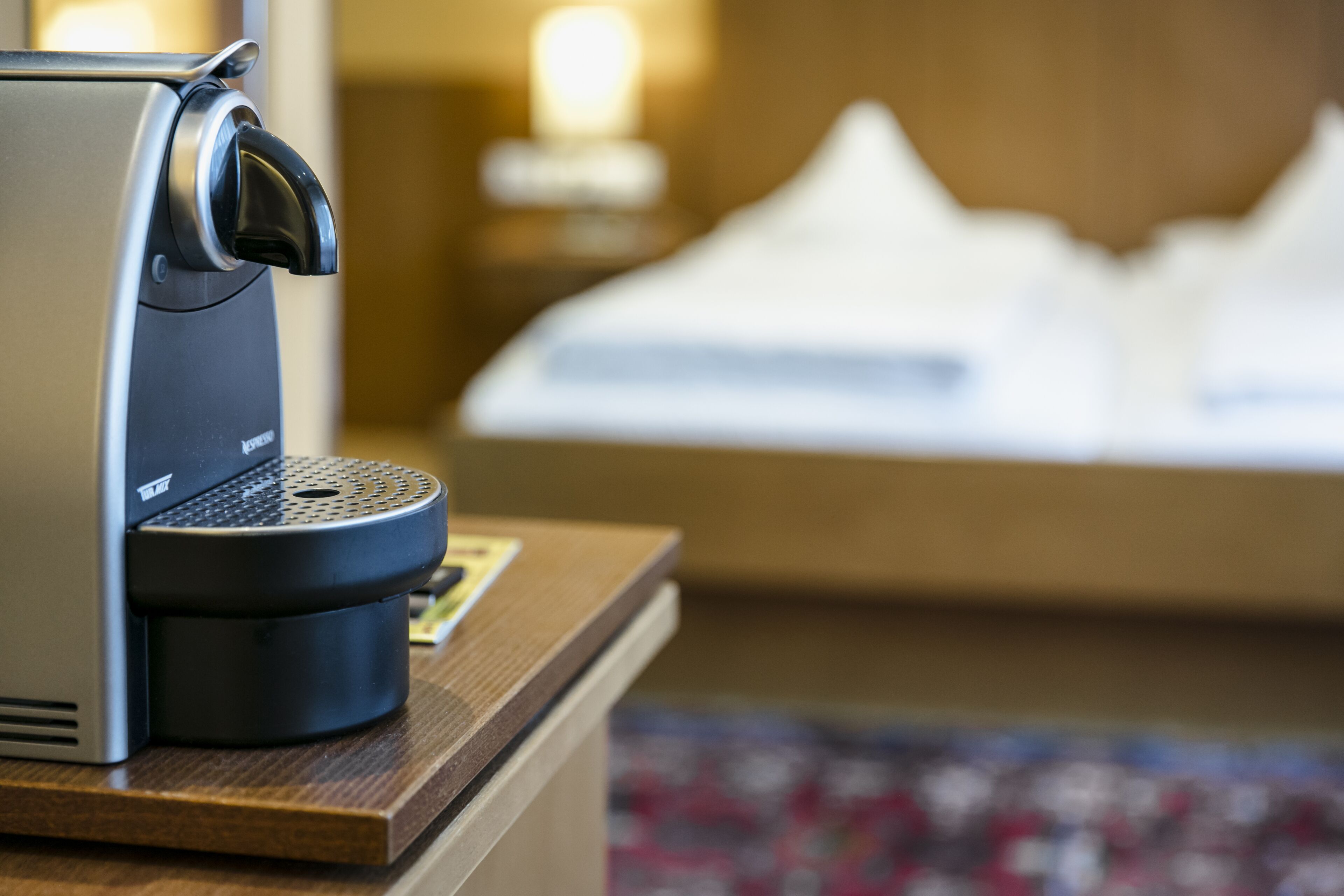 A coffee machine on a wooden counter with a blurred background of a bed and a wall-mounted light.