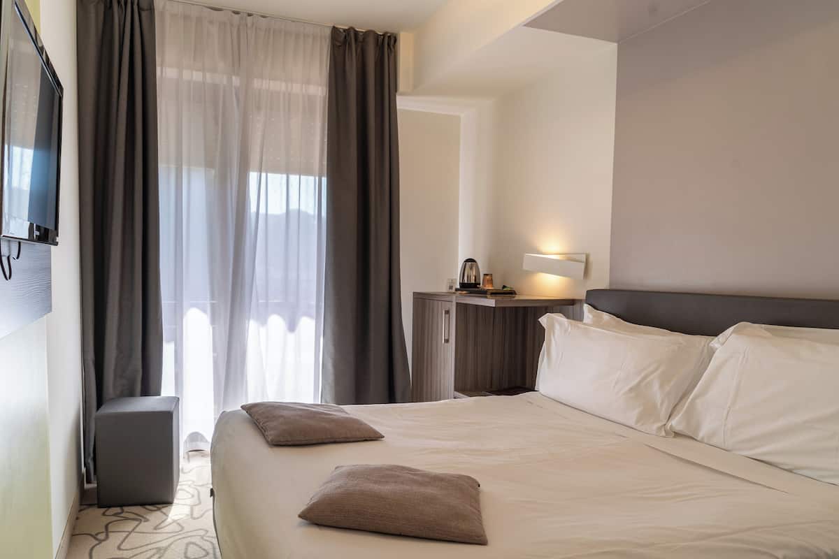 A hotel room with a bed, a bedside table with a lamp, a TV, and a window with curtains.