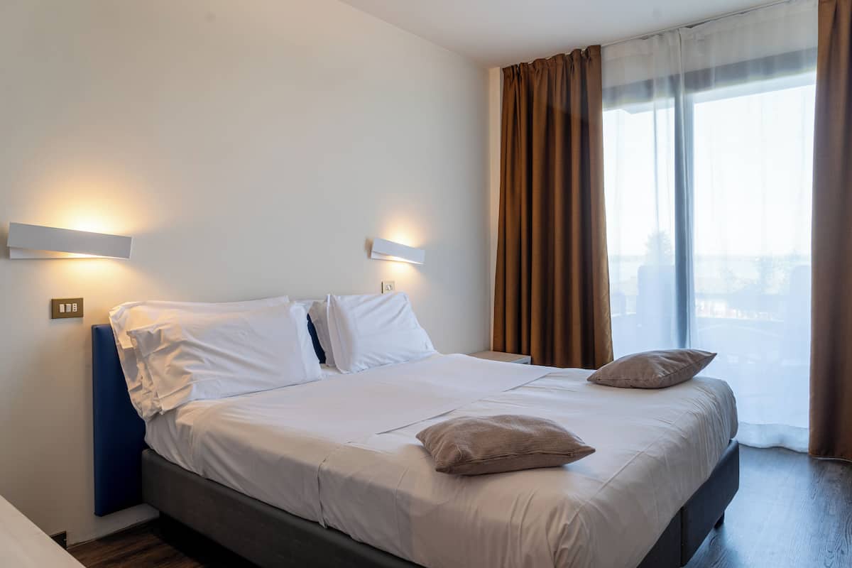 A hotel room with a bed, two bedside lamps, a window with curtains, and a city view.
