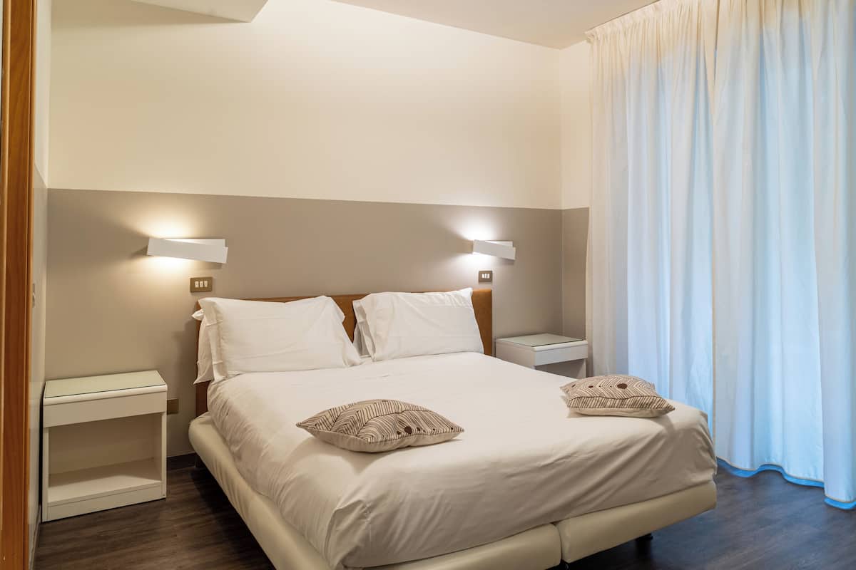 A neatly made bed with two pillows, a bedside table with a lamp, and a window with curtains.