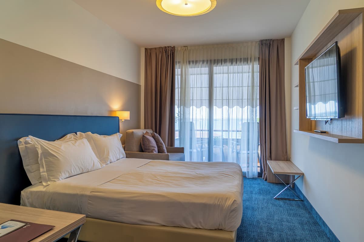 A hotel room with a large bed, a desk, a chair, a TV, and a view of the city through the window.