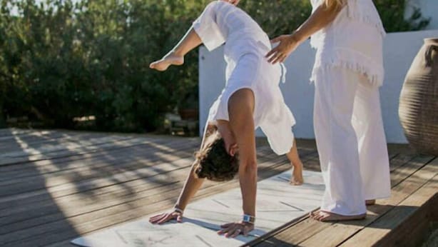 Una persona realiza una postura de yoga en una terraza de madera, con una palmera y un edificio al fondo.