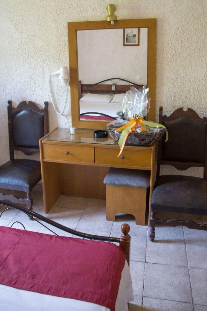 Una habitación con un escritorio de madera, dos sillas, una cama con una colcha roja, un espejo y una mesita de noche con una lámpara.