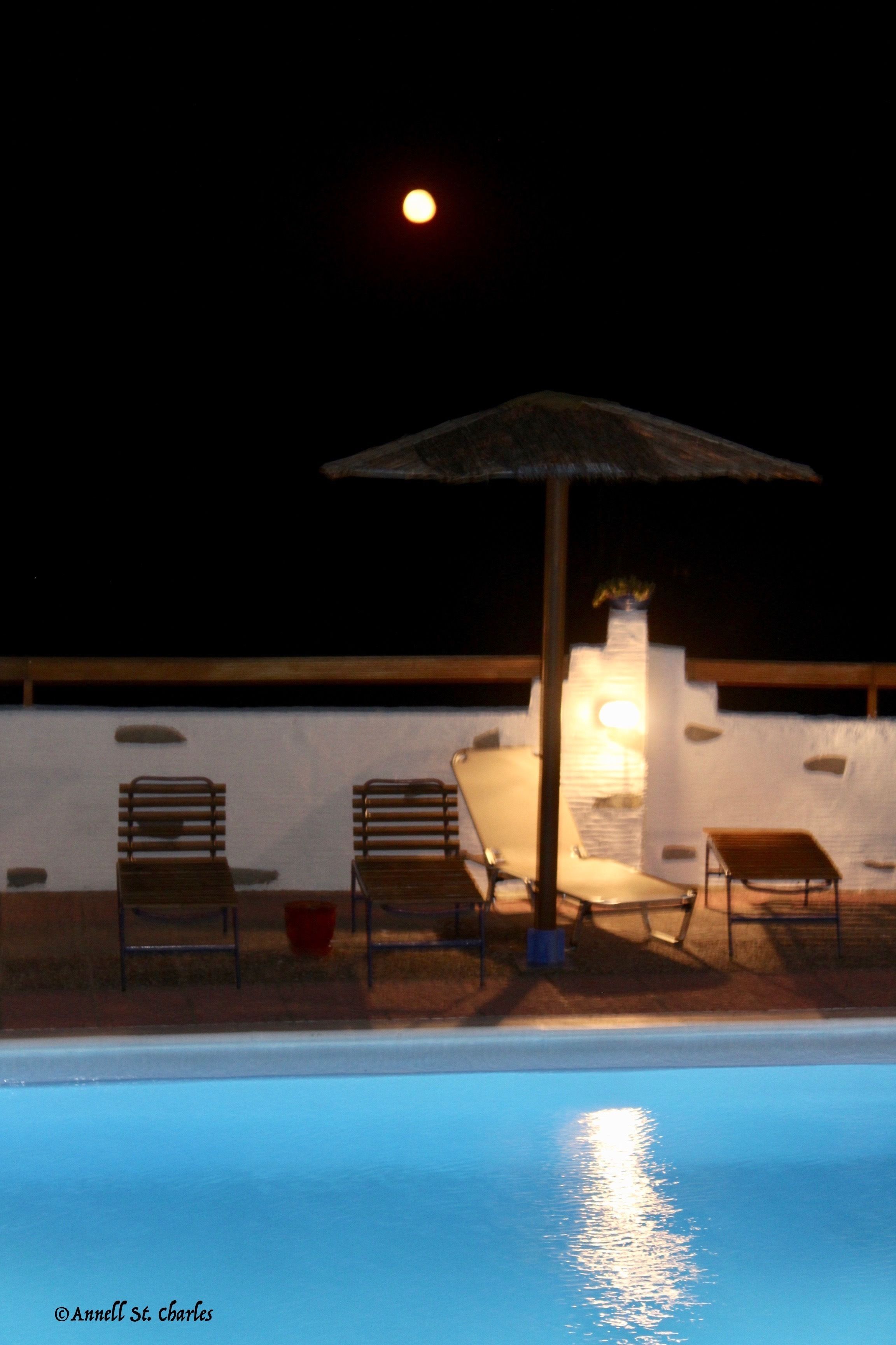 A poolside area with lounge chairs and an umbrella, a white wall, and a moon in the background.