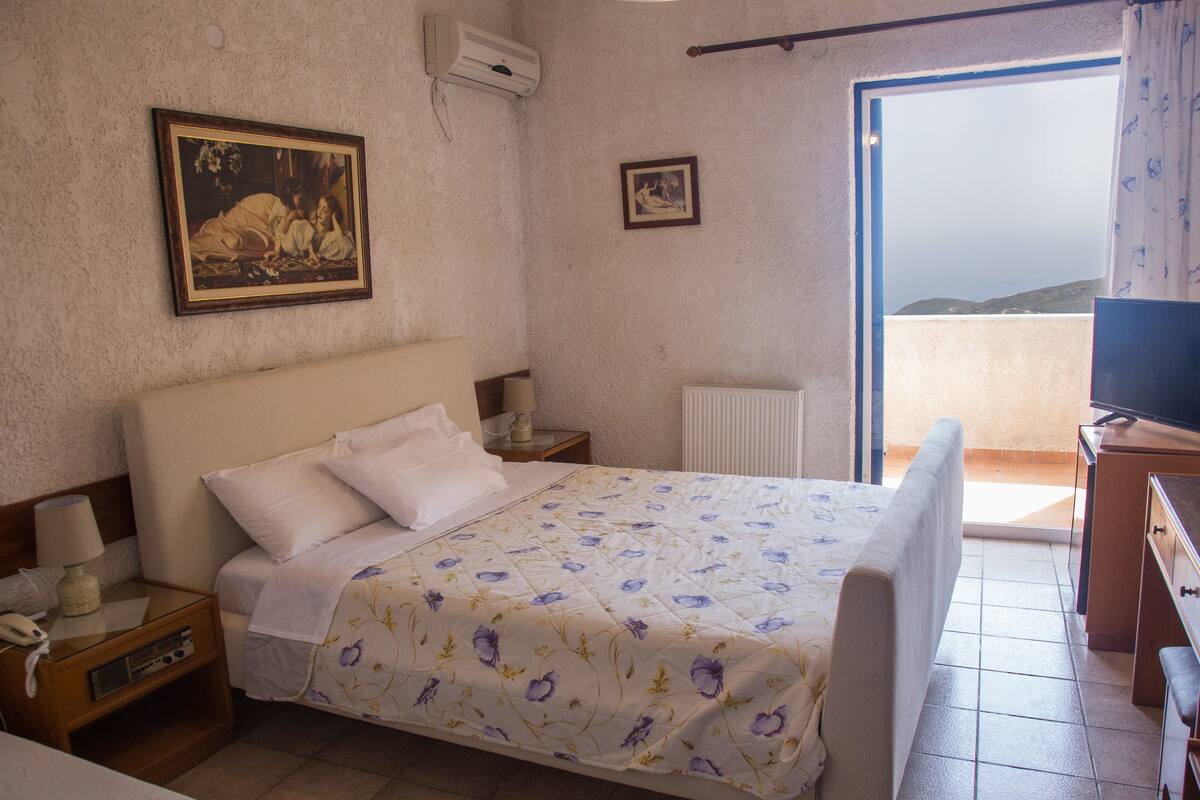 Quarto de hotel com uma cama, mesa de cabeceira com abajur, um quadro na parede e vista pela janela.
