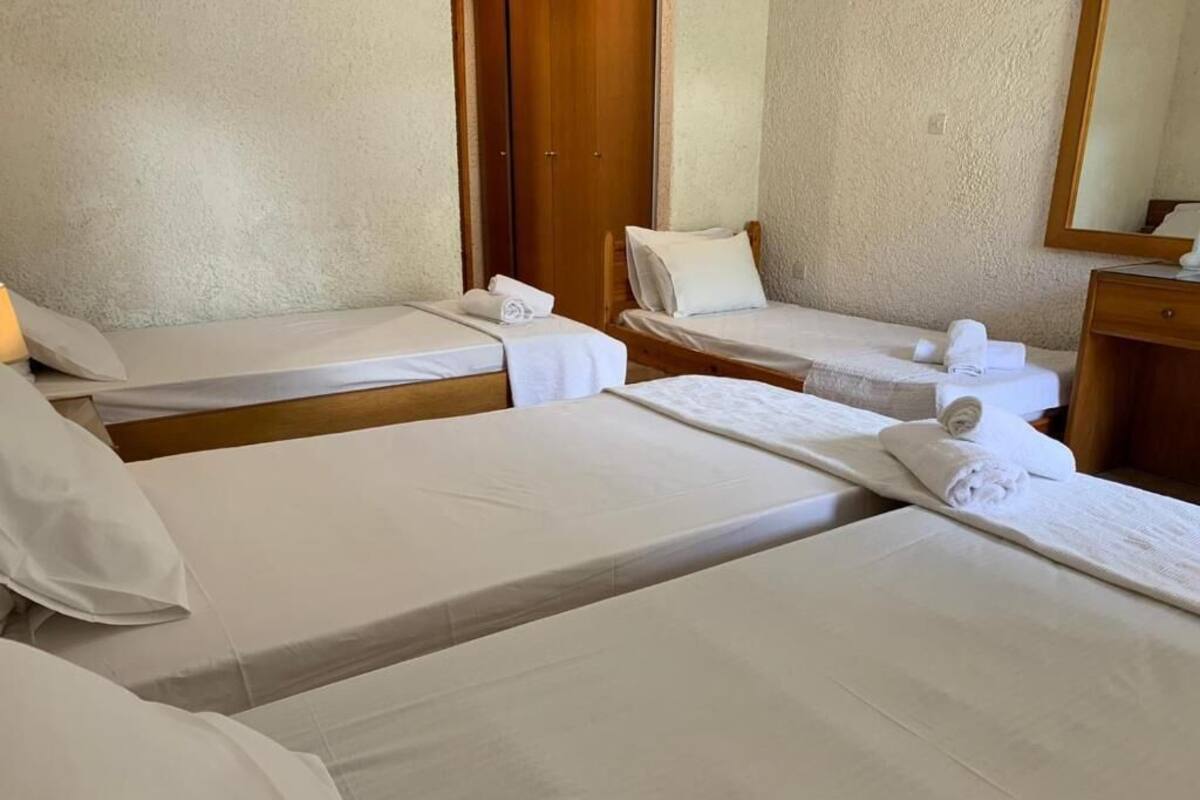 Quarto de hotel com três camas de solteiro, cada uma com colchão e travesseiro branco, e um guarda-roupa de madeira ao fundo.