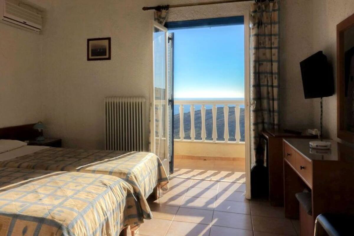 Quarto de hotel com duas camas, radiador, escrivaninha, televisão e varanda com vista para o mar.
