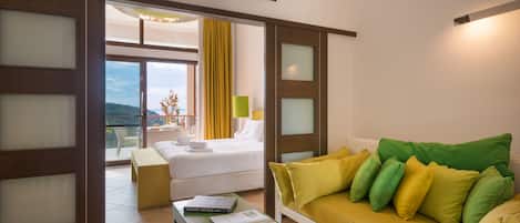 Habitación de hotel con cama, sofá verde con cojines amarillos y verdes, una mesita con un libro, y vistas al exterior a través de un ventanal grande.