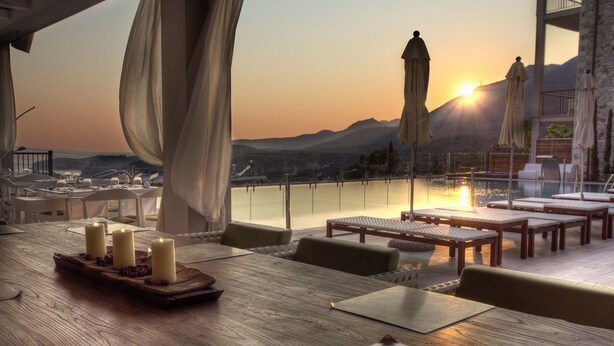 A dining area with a wooden table set for four, surrounded by chairs, with a view of the sunset over a body of water and mountains in the background. - Salvator Villas & Spa Hotel (Parga)