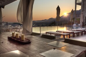A dining area with a wooden table set for four, surrounded by chairs, with a view of the sunset over a body of water and mountains in the background. - Salvator Villas & Spa Hotel (Parga)