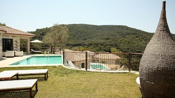 Un área de piscina con sillones de descanso, una terraza cercada y vistas a una colina boscosa.