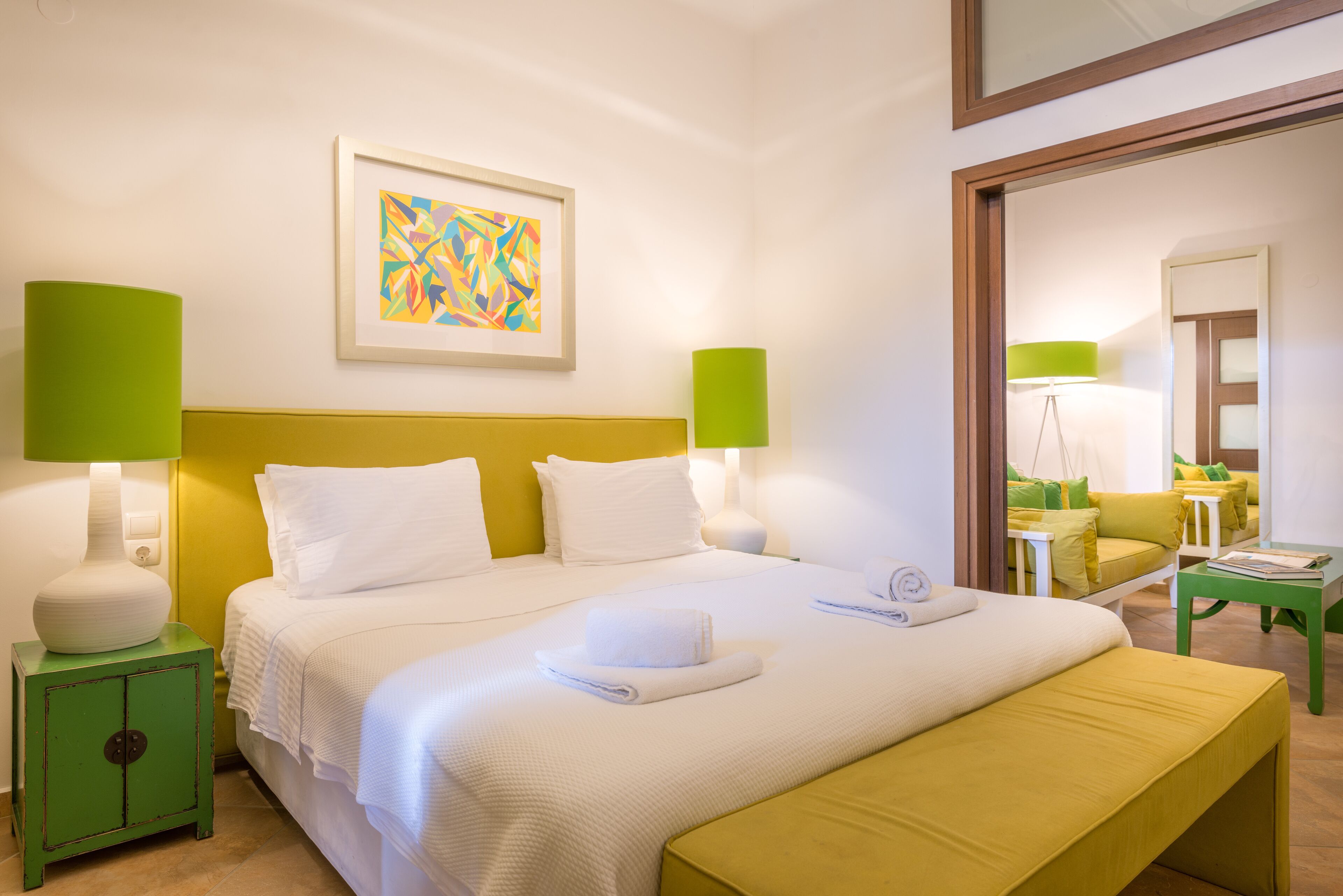 Ein Hotelzimmer mit einem Bett, Nachttischen in Grün, einer grünen Bank, einem Gemälde an der Wand und einem Spiegel.