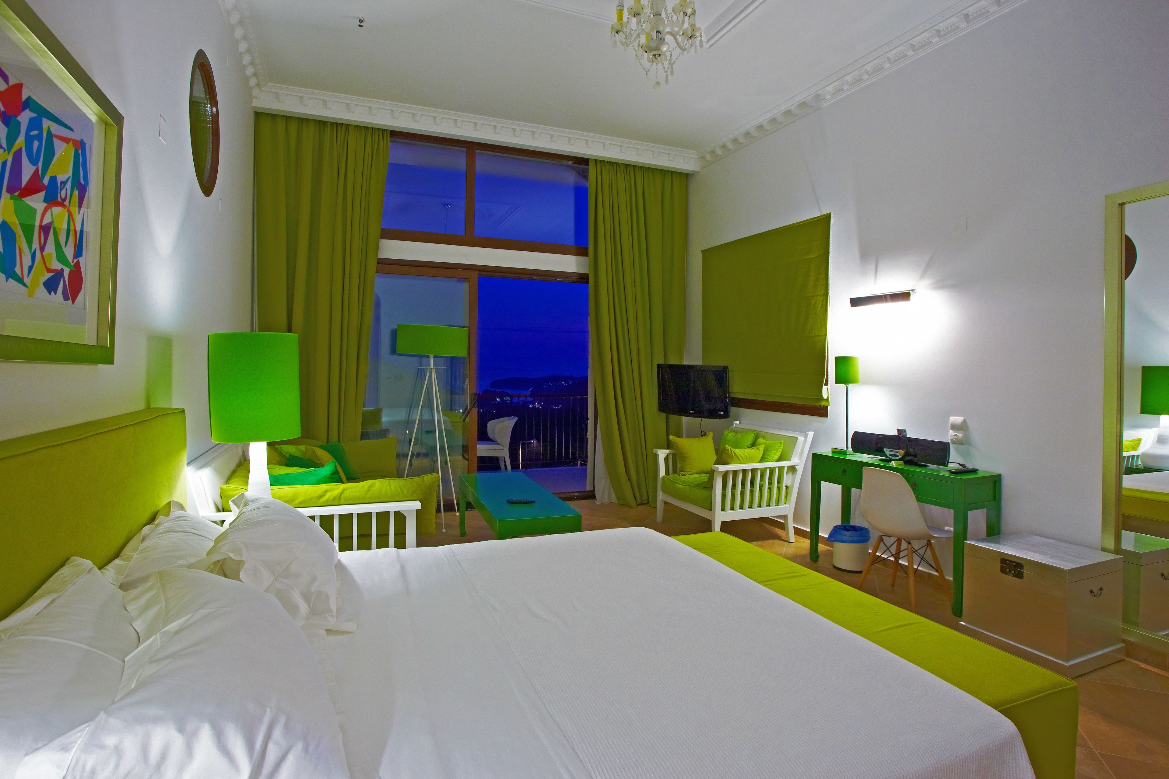 Habitación de hotel con una cama grande, un escritorio, una silla, un televisor y vistas a la ciudad de noche.