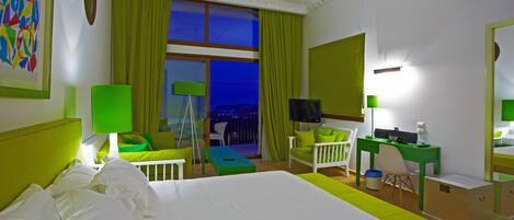 Quarto de hotel com uma cama grande, uma mesa, uma cadeira, televisão e vista da cidade à noite.