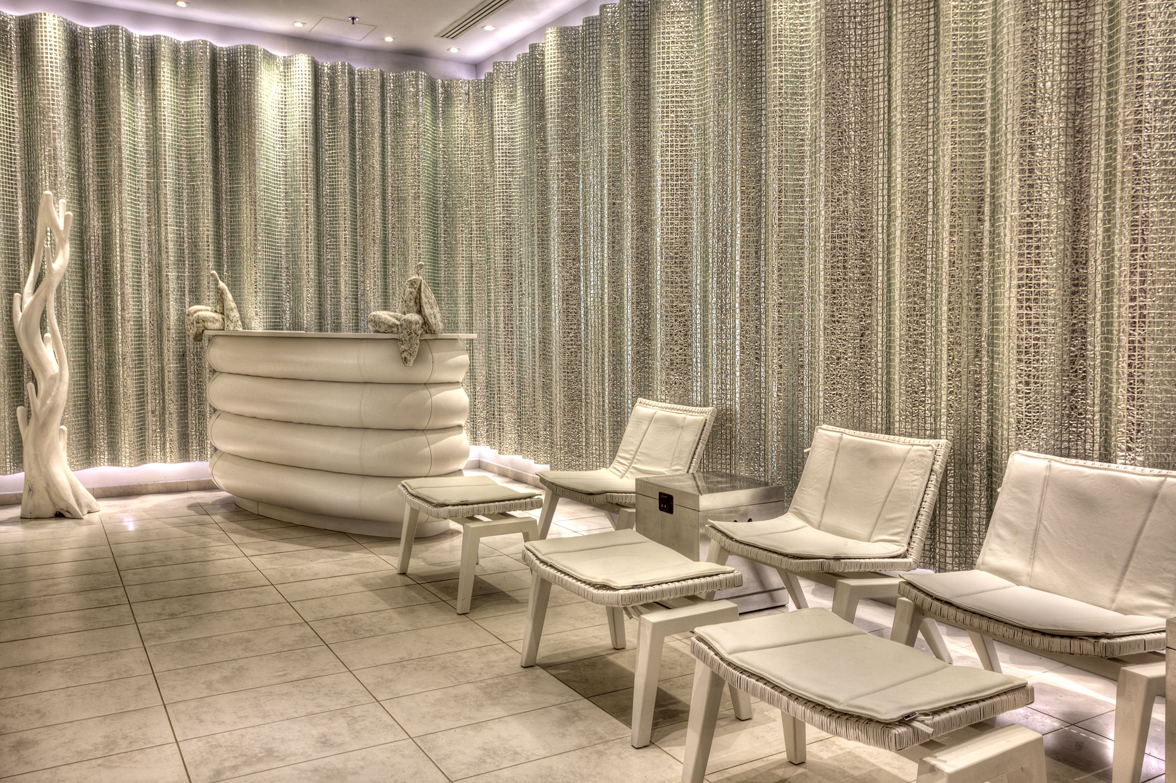 A luxurious spa area with a large, round, white bathtub, several white cushioned chairs, and a textured wall backdrop.