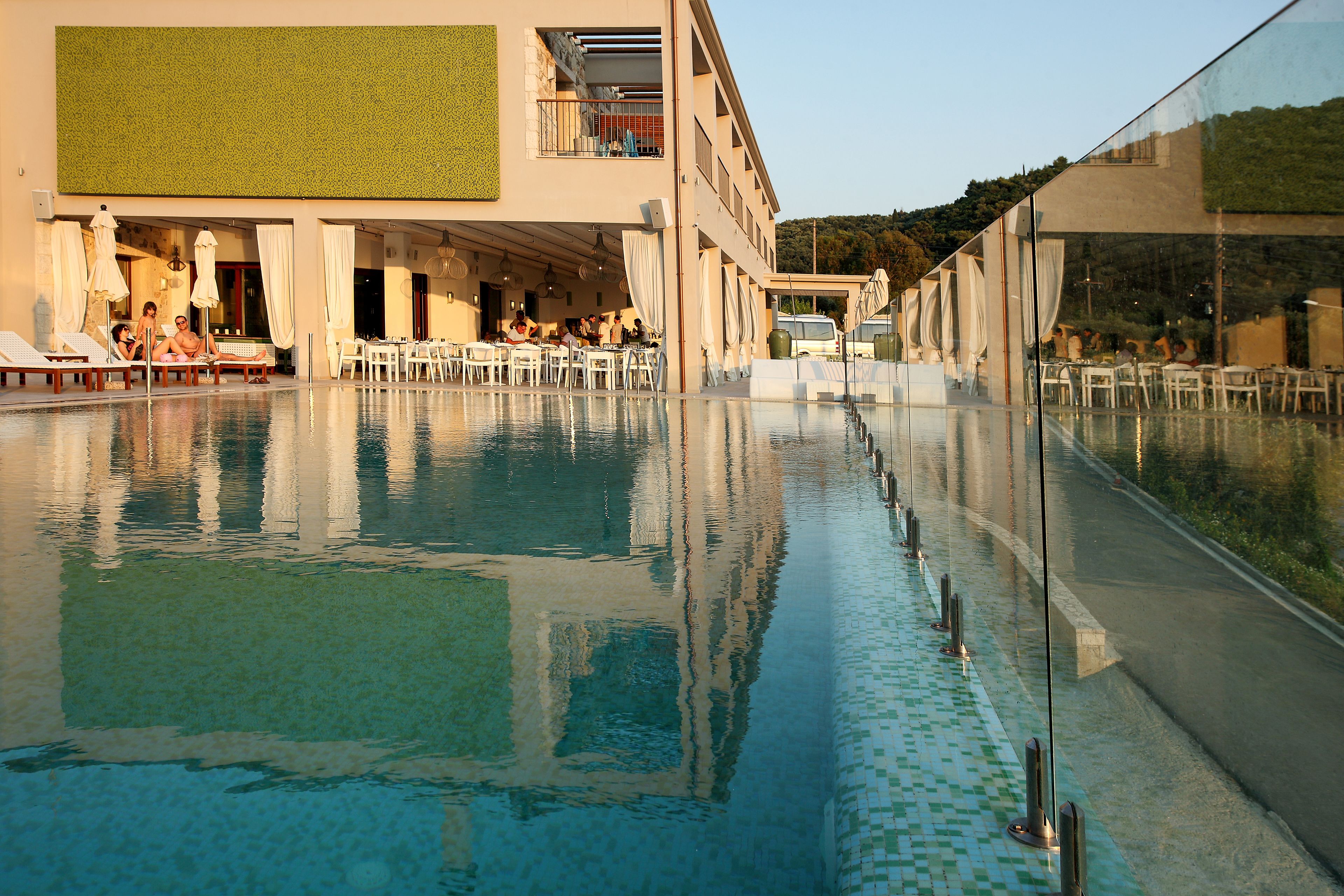 An infinity pool with clear water, surrounded by white lounge chairs and tables, and a building with a green wall and balcony overlooking the pool.