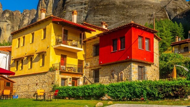 A building with a stone facade and a red roof, situated in front of a large rock formation. - Archontiko Mesohori (Kalabaka)