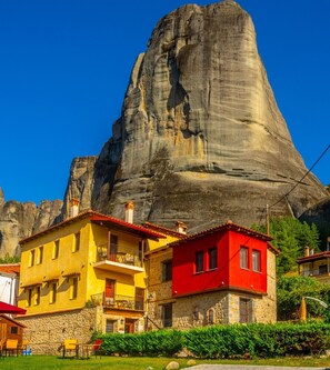 A building with a stone facade and a red roof, situated in front of a large rock formation. - Archontiko Mesohori (Kalabaka)