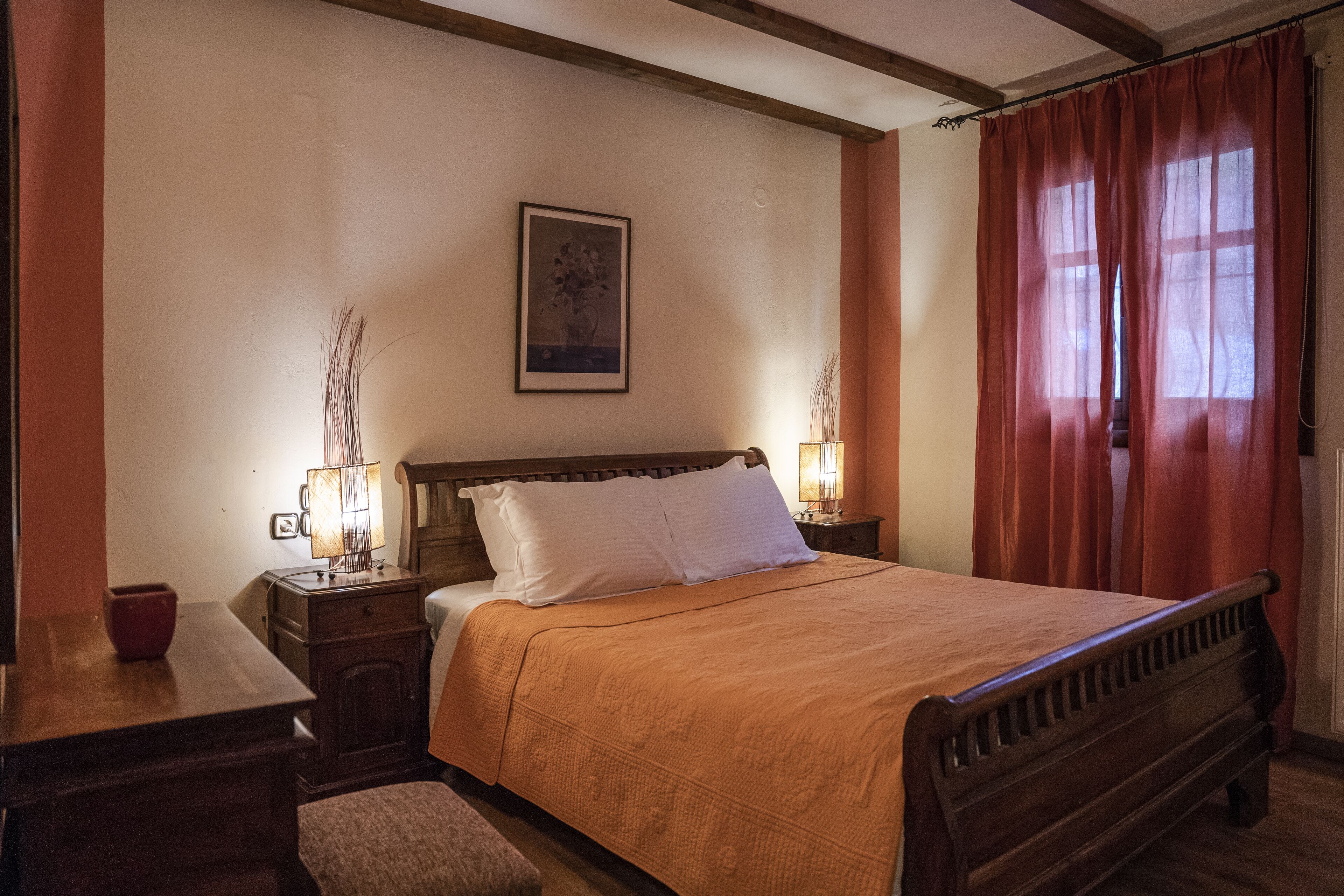 A neatly arranged bedroom with a wooden bed, two bedside tables with lamps, a small table with a red bowl, and a framed picture on the wall.
