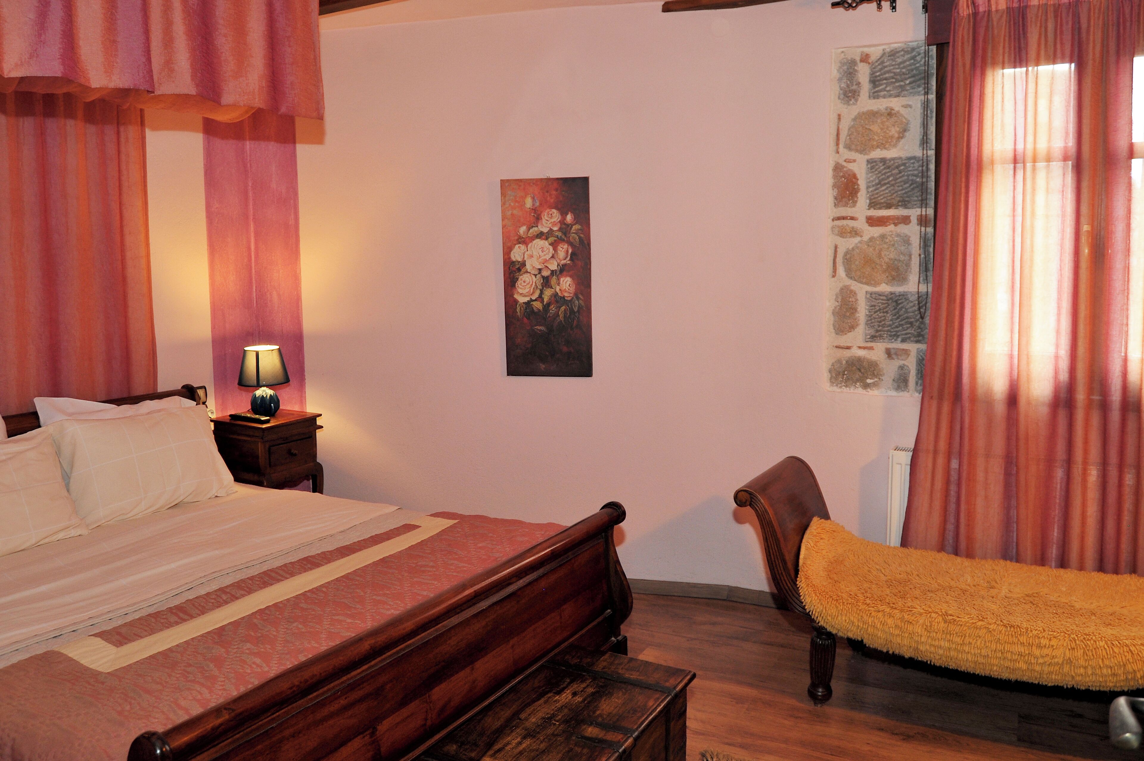 A bedroom with a wooden bed, a nightstand with a lamp, a chair with a yellow cushion, and a painting on the wall.