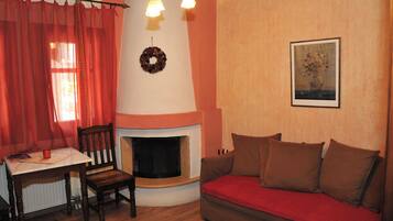 Ein gemütliches Wohnzimmer mit Kamin, einer Couch, einem Tisch mit einem Buch und einem Fenster mit roten Vorhängen.