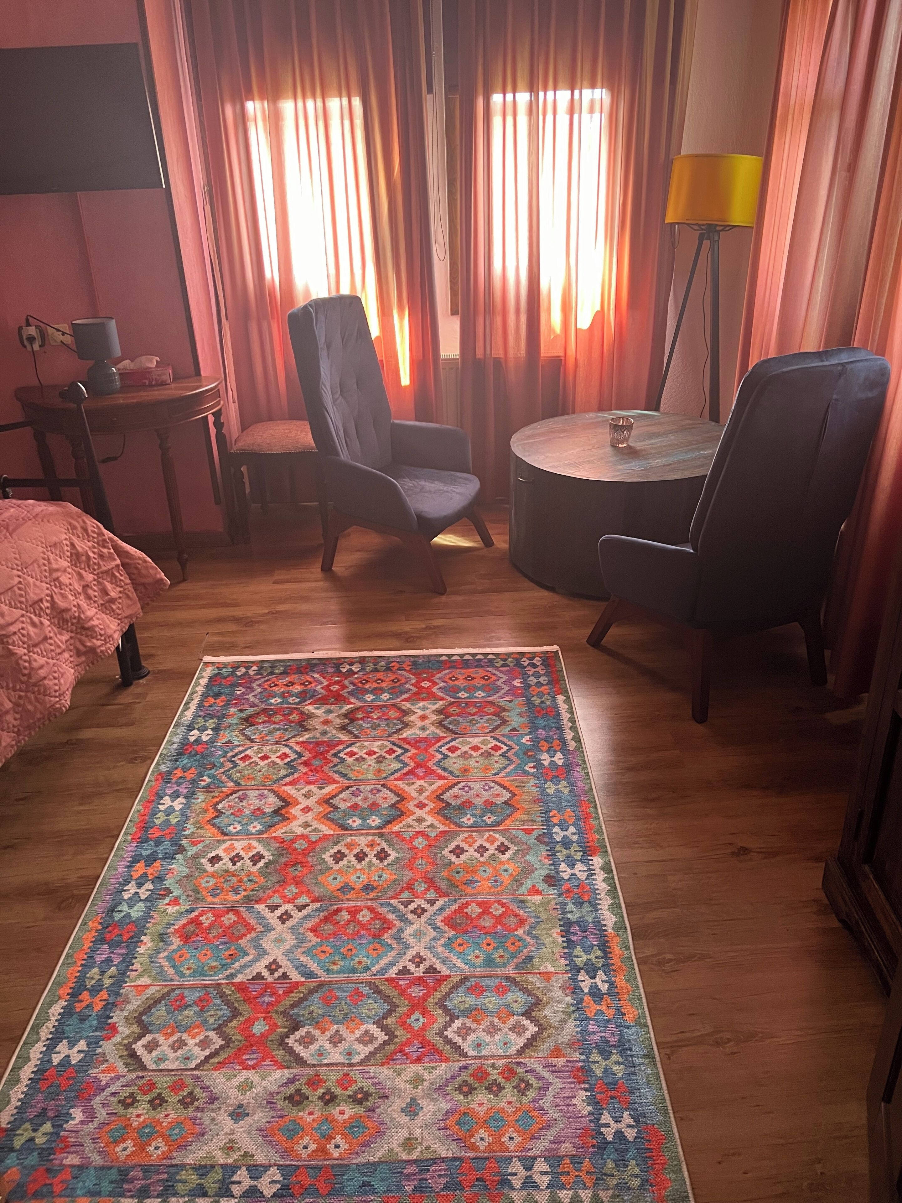 Une pièce avec un tapis à motifs, deux chaises, une petite table avec une lampe et un lit recouvert d’une couverture rose.