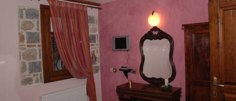 Una habitación con paredes rosadas, una ventana con marco de piedra, un aparador de madera, un espejo, un televisor pequeño y una puerta.