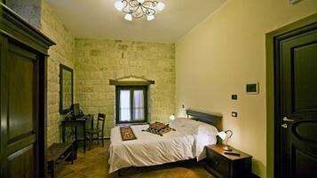 Una camera da letto con parete in pietra, un letto con copripiumino bianco, una scrivania con un computer, una sedia e un comodino con una lampada.