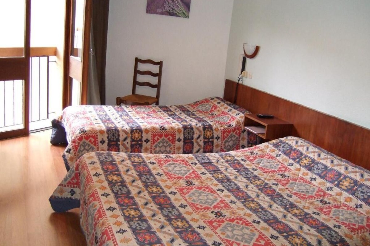 A room with two beds, a chair, a wooden headboard, a picture on the wall, and a window with curtains.