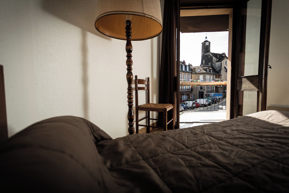 A bedroom with a bed, a wooden chair, and a lamp. A window reveals a street view with buildings and a tower.