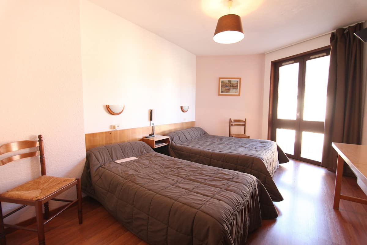 Ein Zimmer mit zwei Einzelbetten, einem Holzstuhl, einem Schreibtisch und einem Fenster mit Vorhängen.