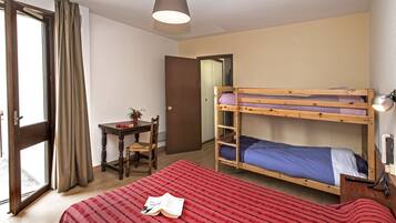 A dormitory room with a bunk bed, a desk with a chair, a small table with a plant, and a door leading to another room.