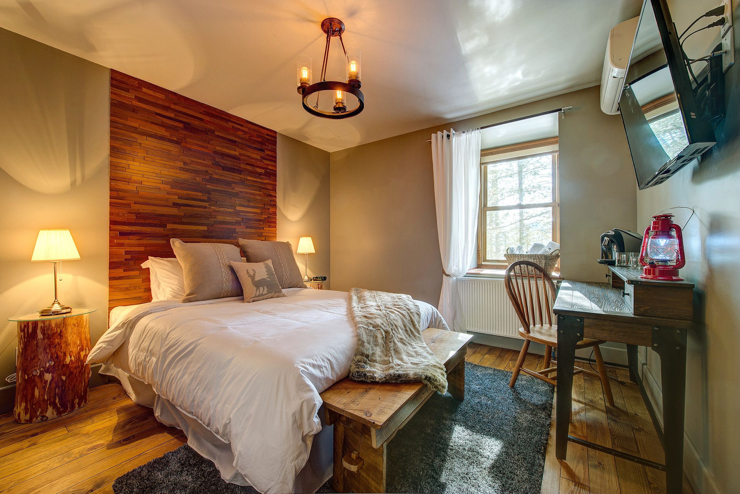 A bedroom with a wooden headboard, a bed with white linens, a bench at the foot of the bed, a desk with a chair, a television mounted on the wall, and a window with curtains.